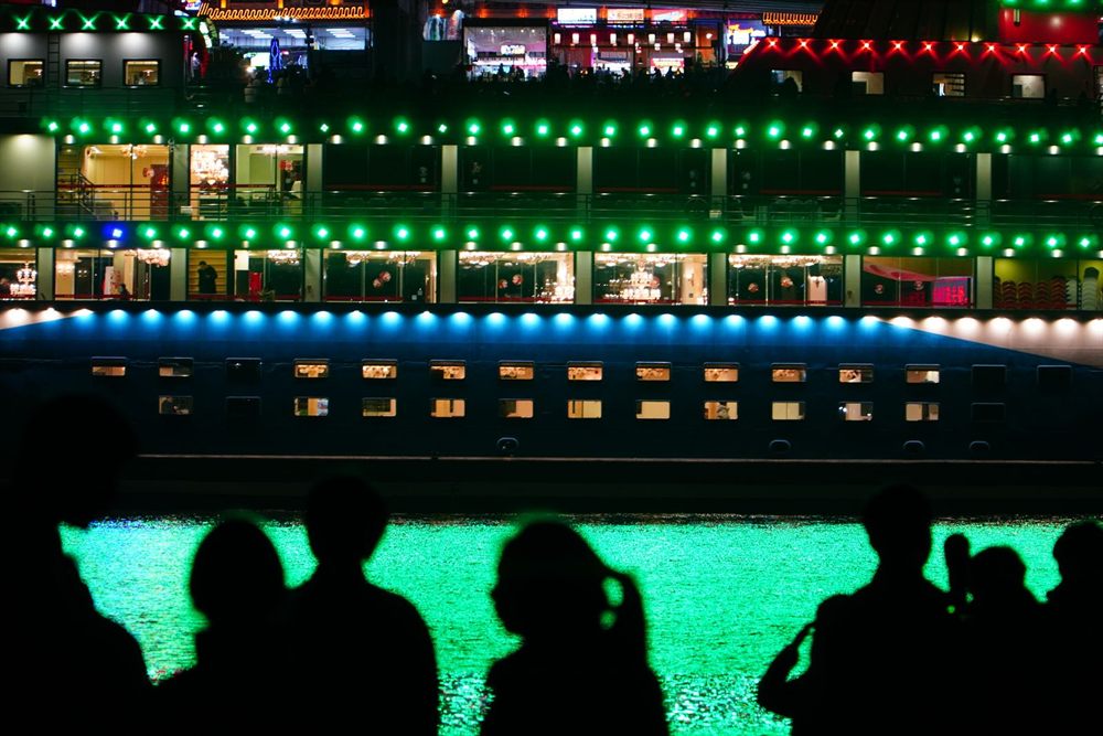 Silhouetted people watching a brightly lit cruise ship at night